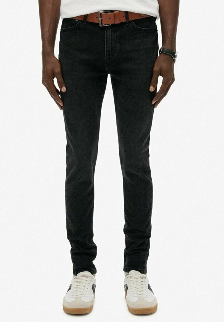 Vaqueros de mezclilla negra con corte slim, cinco bolsillos y un cinturón de cuero marrón. Combinados con zapatillas blancas con acentos negros.