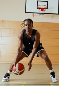 Μαύρη μπλούζα μπάσκετ και σορτς, αθλητική σιλουέτα, επικεντρωμένος στην ντρίμπλα μιας κόκκινης και λευκής μπάλας μπάσκετ, με φόντο ξύλινο γυμναστήριο.