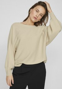 Beżowy sweter w prążki z szerokim dekoltem i długimi rękawami, noszony na czarnych spodniach, o luźnym kroju i miękkiej teksturze.