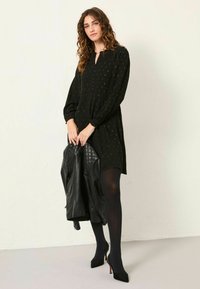 Schwarzes Kleid mit V-Ausschnitt, langen Ärmeln und goldenen Punkten. Kombiniert mit schwarzen Strumpfhosen und Stiletto-Absätzen, hält sie eine schwarze, gesteppten Lederjacke.