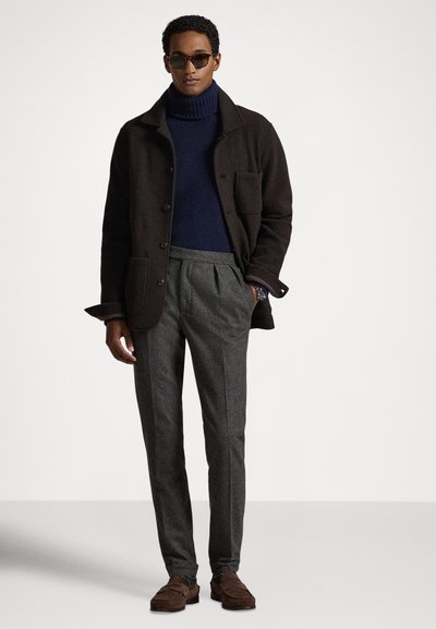 Tumši brūns vilnas mētelis ar apkakli un kabatām, tumši zils augstas apkakles džemperis, pelēkas piegrieztas bikses un brūnas zamšādas loafers kurpes. Uzliktas melnas saulesbrilles.