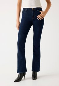 Jean flare bleu foncé en denim, avec une taille mi-haute, un design à cinq poches et des détails de couture subtils. Porté avec des talons noirs.