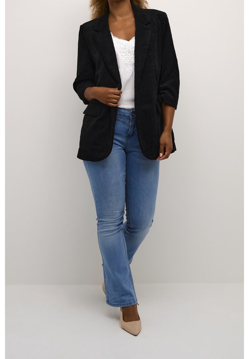 Personne vêtue d'un blazer noir, d'un haut blanc orné de dentelle, d'un jean bleu clair et de talons pointus beiges, debout devant un fond uni.