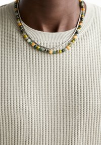 Gerippter, hellgrüner Pullover mit Rundhalsausschnitt, versehen mit zwei geschichteten Halsketten: einer silbernen Kette und einer farbigen Perlenkette.