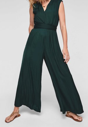 Kobieta ubrana w ciemnozielony, szerokoskalowy kombinezon bez rękawów z dekoltem w kształcie litery V oraz beżowe płaskie sandały, stojąca z nogami rozstawionymi.