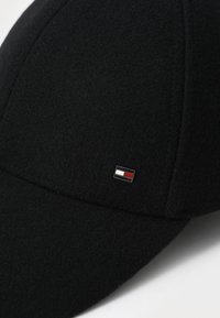 Casquette de baseball en laine noire avec un petit logo en métal rouge, blanc et bleu sur le côté, un bord incurvé et des détails cousus.