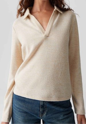 Femme portant un pull beige à manches longues et col en V ainsi qu'un jean bleu, cadrée du cou aux hanches.