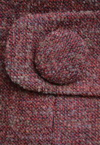 Tkanina wełniana o teksturowanym wzorze z czerwonych, pomarańczowych i szarych włókien. Posiada okrągły, teksturowany guzik z lekko puszystą powierzchnią.