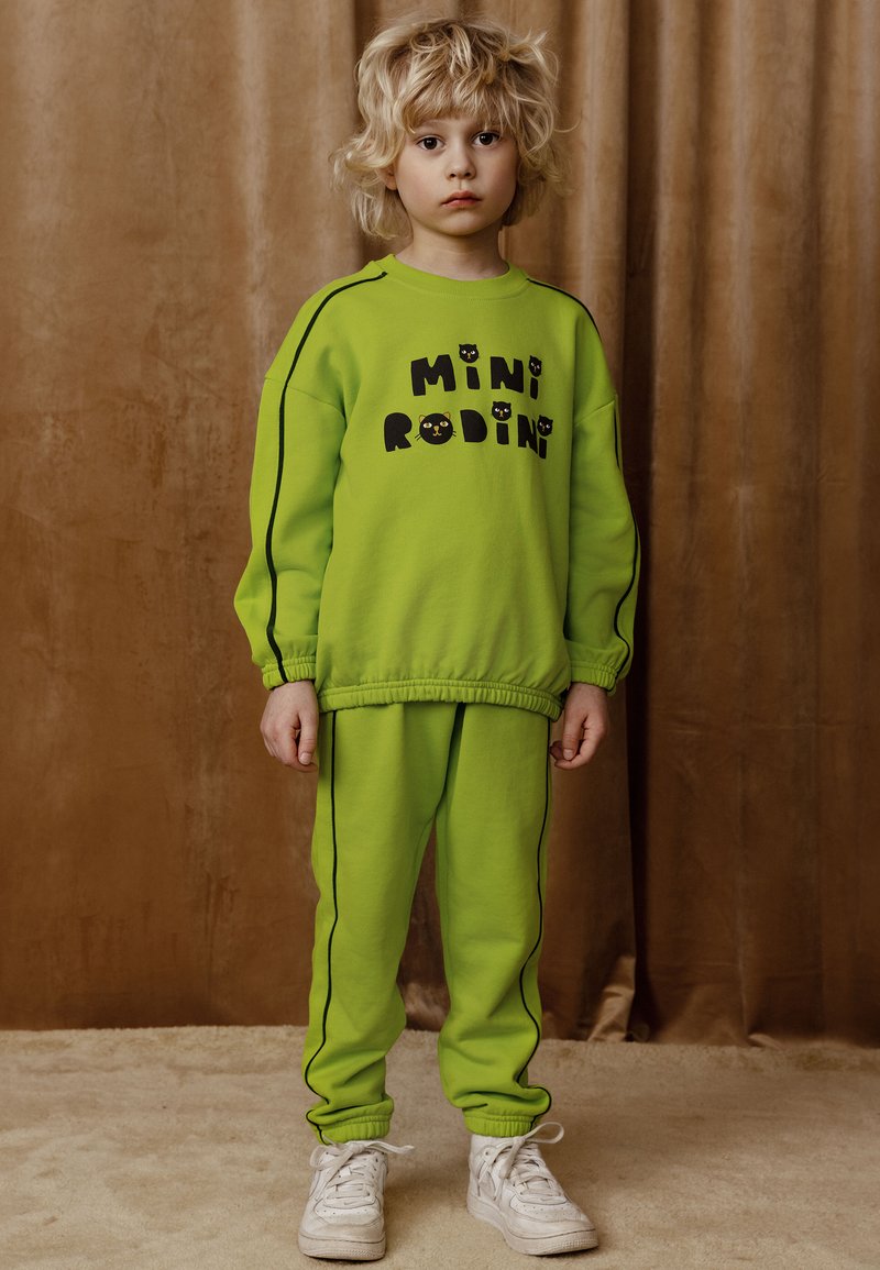 Svetlozelený teplákový komplet s čiernymi akcentmi a logom "MINI RODINI." Dizajn ponúka voľný strih a elastické manžety, skombinovaný s bielymi teniskami.