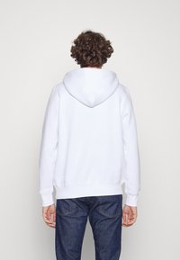 Sudadera blanca con bolsillo canguro y capucha ajustable con cordón. El tejido tiene un aspecto suave y agradable, combinado con jeans de un azul oscuro.