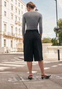 Hall halli pika varrukaga pluus ümmarguse kaelusega, mida kantakse koos mustade põlveni ulatuvate pükste ja mustade paeltega kingadega, vaadatuna tagantpoolt.