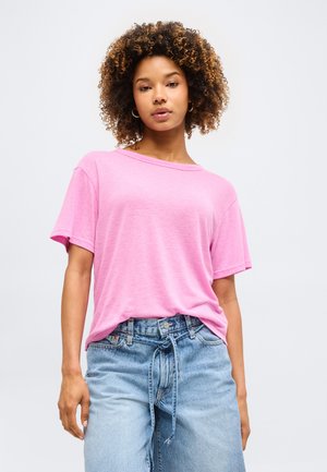 Γυναίκα με σγουρά μαλλιά φοράει ροζ φαρδιά μπλούζα που είναι στραμμένη μέσα σε ψηλόμεσο φως μπλε τζιν με ζώνη δεμένη, απέναντι σε μονόχρωμο φόντο.