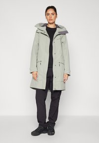 Giacca impermeabile verde chiaro con cappuccio, due tasche frontali e dettagli neri. Indossata sopra una maglia nera e pantaloni scuri. Calzature da esterno.