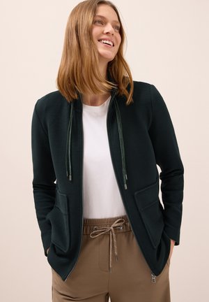Giacca verde scuro con zip in tessuto texture, dettagli con cordoncino e tasche laterali, indossata sopra una camicia bianca e pantaloni beige.