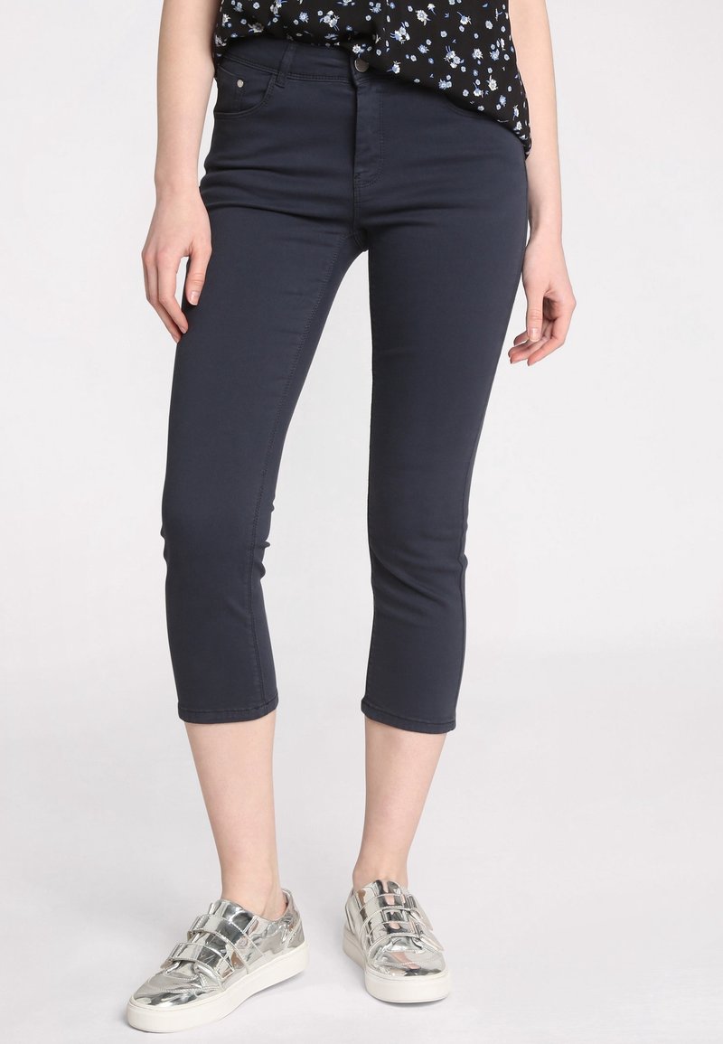 Pantalons cropped bleu marine en tissu stretch avec une coupe ajustée, dotés d'une fermeture à bouton et de poches discrètes. Associés à des baskets argentées.