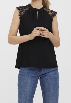 Ærmeløs sort top med blonde-kappelærre og en lille slidse foran, båret med blå denimjeans, hænderne foldet foran.