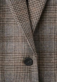 Brązowa marynarka w kratę z teksturowanej mieszanki wełny, z wyciętymi klapami i czarnym przyciskiem jako akcentem. Zbliżenie na szczegóły tkaniny i wzoru.
