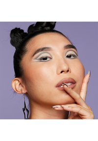 Modèle avec des cheveux tressés et des yeux maquillés en gris avec des motifs géométriques. Ongles longs remarquables avec du vernis noir et nude, sur un fond violet.