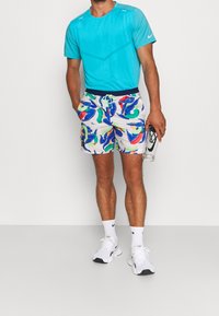 Tyrkysové sportovní tričko, vzorované bílé šortky s modrými a zelenými designy, bílé tenisky a průhledná láhev na vodu.