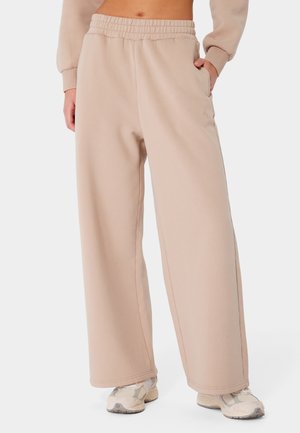 Beżowe spodnie o szerokich nogawkach z elastycznym pasem, boczne kieszenie, noszone z jasnymi trampkami i dopasowanym długim rękawem odsłaniającym brzuch.