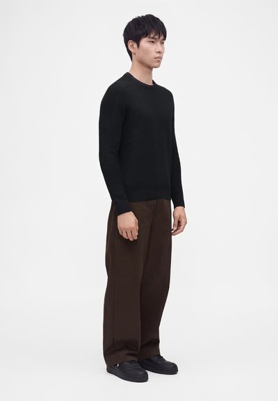 Jeune homme debout de profil portant un pull noir à manches longues, un pantalon marron à jambes larges et des chaussures noires sur un fond blanc uni.