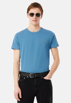 Moški, ki nosi modro majico s kratkimi rokavi, črnim pasom, črnimi kavbojkami, sončnimi očali in srebrno zapestnico, z roko v žepu, na belem ozadju.