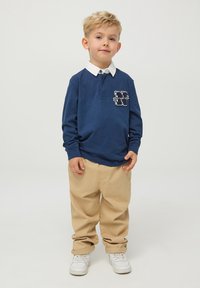 Niebieska koszulka polo z długim rękawem, białym kołnierzykiem i logo na piersi, w komplecie z beżowymi spodniami i białymi trampkami, na neutralnym tle.