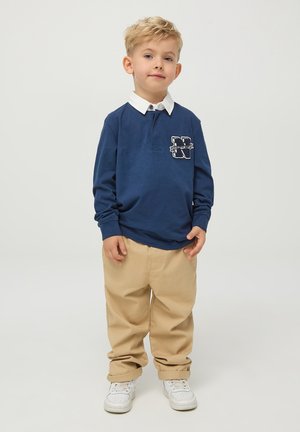 Polo shirt blu a maniche lunghe con colletto bianco e logo sul petto, abbinato a pantaloni beige e sneakers bianche, sfondo neutro.