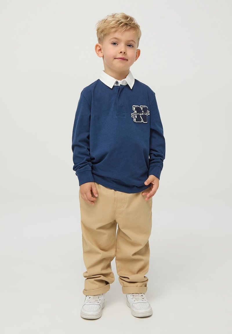 Niebieska koszulka polo z długim rękawem, białym kołnierzykiem i logo na piersi, w komplecie z beżowymi spodniami i białymi trampkami, na neutralnym tle.