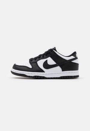 Baskets basses noires et blanches Nike avec swoosh noir, lacets noirs et semelle intermédiaire blanche sur un fond gris clair uni.