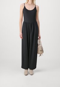 Mono negro sin mangas con un torso ajustado y texturizado y pantalones de pierna ancha. Combinado con un bolso de mano beige claro y botas hasta el tobillo.