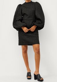 Schwarzes, strukturiertes Minikleid mit Puffärmeln und hohem Ausschnitt. Kombiniert mit schwarzen, offenen Mules mit Blockabsatz.