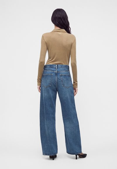 Femme debout, de dos, portant un haut à manches longues beige, un jean bleu pattes larges et des chaussures à talons noires sur fond blanc.
