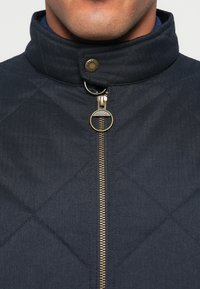 Casaco acolchoado azul com gola alta, apresentando um fecho dourado e um detalhe metálico. A textura da superfície é lisa com um padrão espinha de peixe.