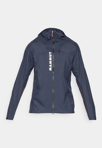 Marineblauwe lichtgewicht jas met een rits aan de voorkant en een capuchon, voorzien van witte "MAMMUT"-tekst en zwarte accenten op de mouwen en zijkanten.