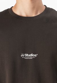 Camiseta de algodón de cuello redondo en marrón oscuro con logotipo impreso en blanco "JJ Studios" y texto debajo. Diseño sencillo, textura suave.
