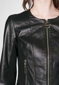 Giacca di pelle nera con colletto rotondo, zip anteriore e tasche laterali. Presenta minuteria dorata e una texture liscia.