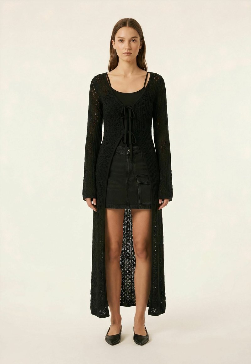Femme portant un long cardigan noir en maille ajourée par-dessus un débardeur noir et une mini-jupe en jean noire avec des ballerines pointues noires, debout droite.