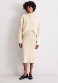 Pull en tricot beige avec un col montant et une coupe ample, associé à une jupe longue assortie. Le mannequin porte des mocassins noirs.