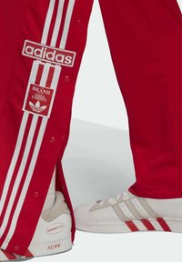 Czerwone spodnie dresowe z białym wzorem trzech pasków i naszywkami z logo, zestawione z białymi i szarymi butami Adidas Superstar.