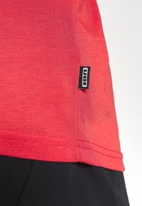 Red cotton T-shirt with short sleeves and a black label at the hem featuring white text. Smooth texture, casual fit.