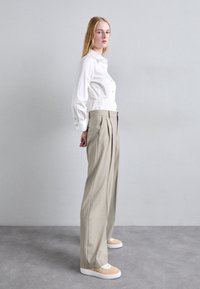 Weißes Button-Up-Hemd, high-waist hellbeige, faltige Hose und gemusterte hellbeige Sneakers mit weißen Sohlen, die vor einem grauen Hintergrund stehen.