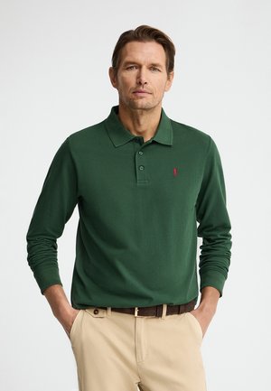 Hombre con una camisa tipo polo de manga larga verde oscuro, pantalones beige y cinturón marrón, de pie con las manos en los bolsillos.