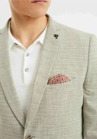 Ljusgrå rutmönstrad kavaj med slag, bar över en texturerad vit polo. En mönstrad orange och röd näsduk är i bröstfickan.