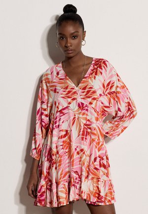 Kobieta o ciemnych włosach związanych w kok, nosząca kolczyki koła oraz sukienkę z dekoltem w serek, długimi rękawami i falbaną na dole, w wzór liści w kolorach różu, pomarańczu i czerwieni.