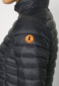 Zwarte pufferjack met een gestikte textuur, hoge kraag en oranje ronde logopatch. De stof lijkt glanzend en lichtgewicht, geschikt voor koud weer.