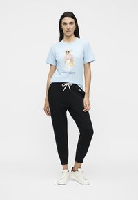 Γυναίκα που φοράει ανοιχτό μπλε μπλουζάκι με σχέδιο αρκούδας, μαύρο παντελόνι με κορδόνι και λευκά αθλητικά παπούτσια, στέκεται μπροστά σε λευκό φόντο.