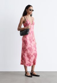 Femme portant une robe midi fleurie rose, des ballerines noires pointues, des lunettes de soleil et portant un sac à main noir sur un fond blanc uni.