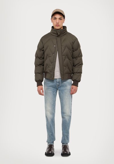 Olive zöld steppelt bomber dzseki magas gallérral, alatta szürke ing, világoskék farmer és sötétbarna bőrcipő.