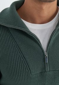 Tmavě zelený svetr s texturovaným žebrovaným vzorem, s uzávěrem na půl zip a širokým límcem. Pod ním je vidět bílý svetřík s kulatým výstřihem.